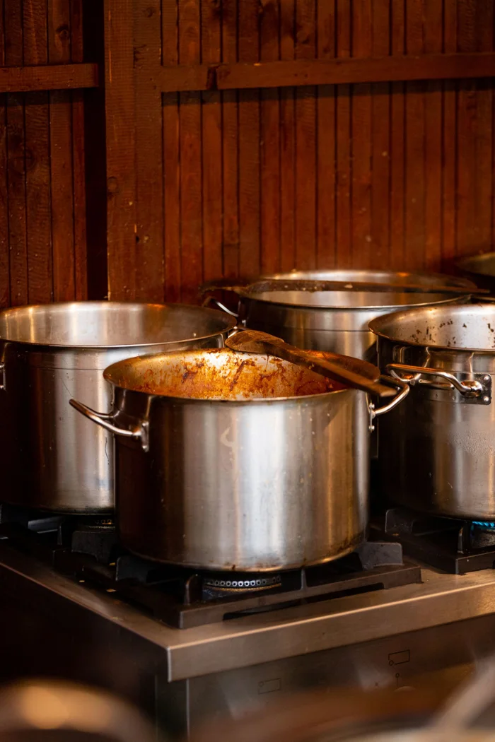Stainless steel cooking pots with wooden spatula on a stove in a restaurant kitchen.