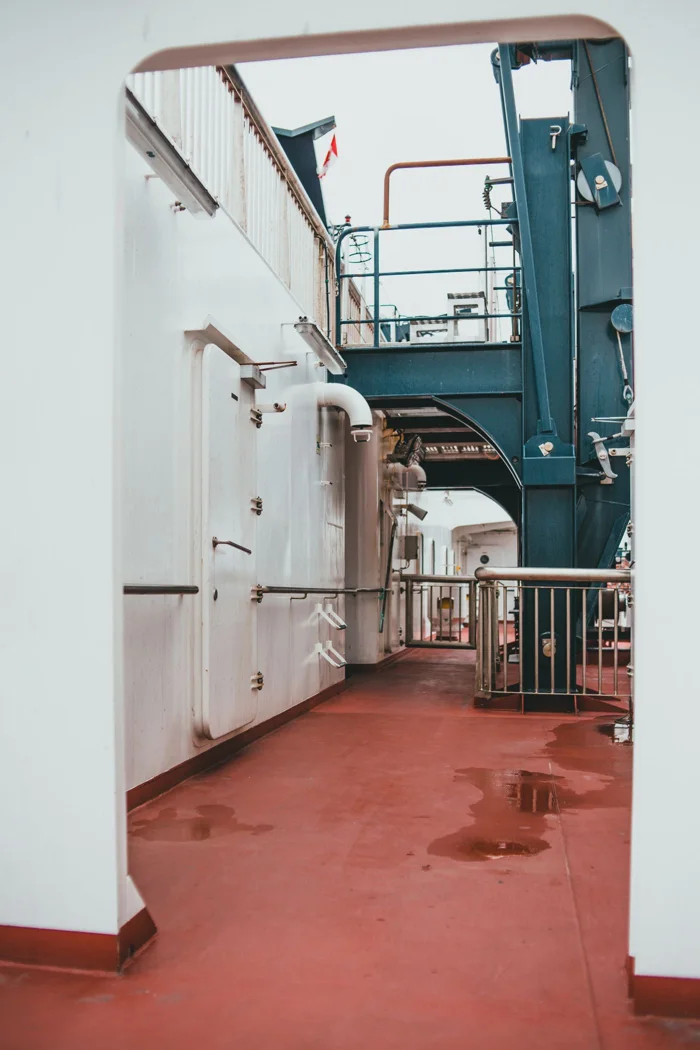 A view of a ship's interior corridor with metal doors and piping under overcast lighting.