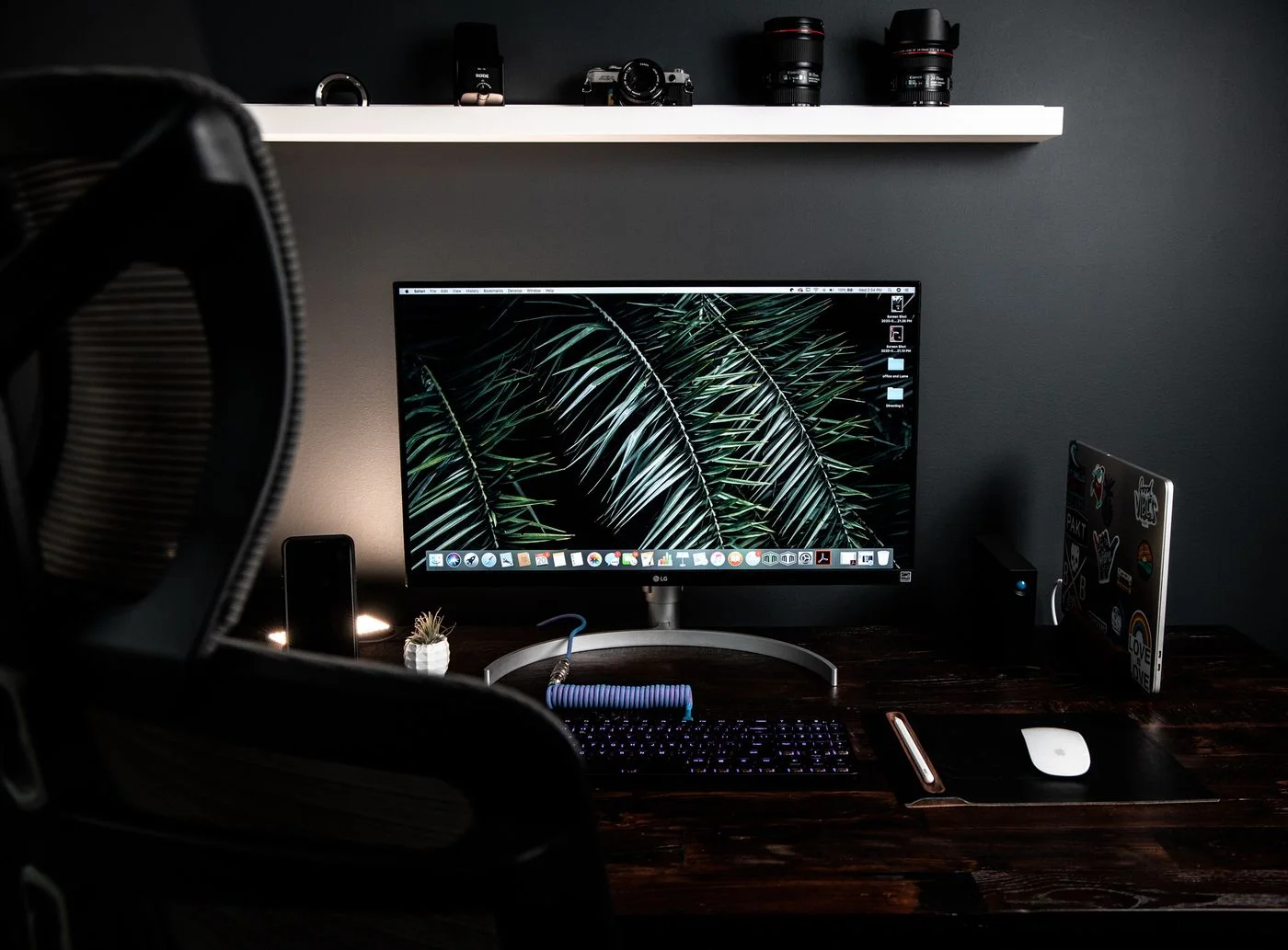 Stationery computer with keyboard and mouse on desk placed under white shelf in dark workspace with armchair near table in modern room.