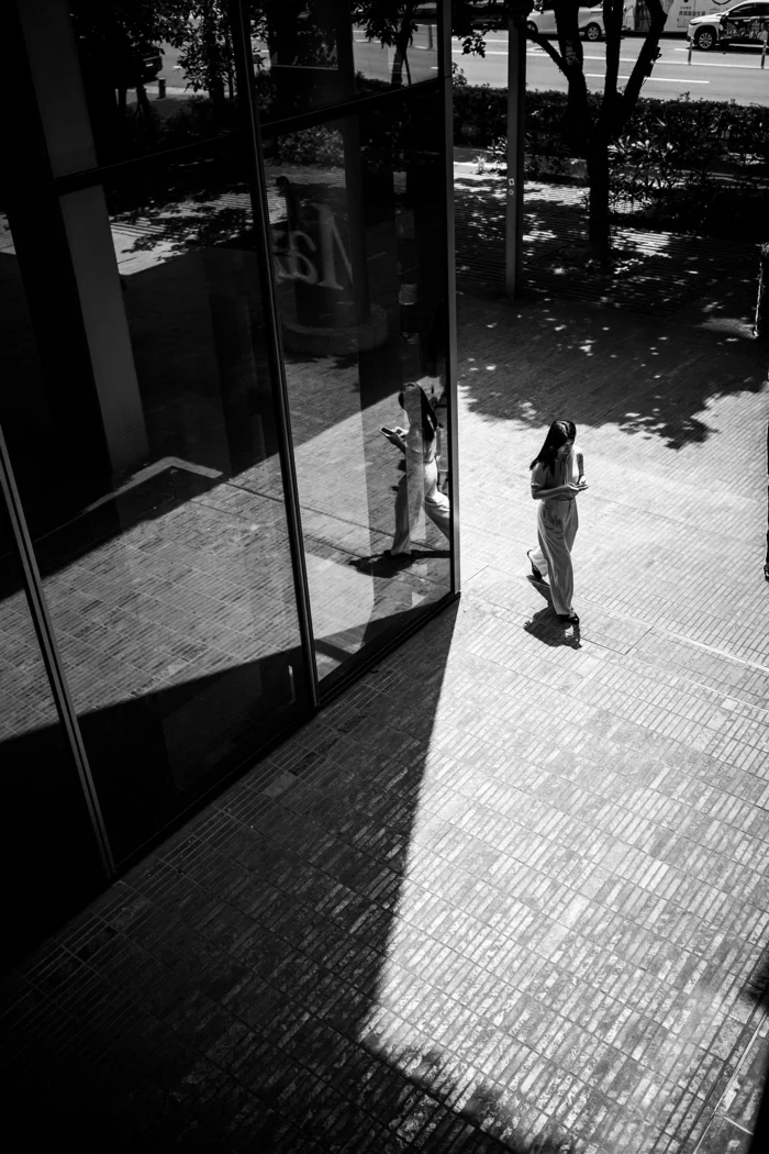 Black and white photo of a woman using a phone, reflected in glass outdoors.