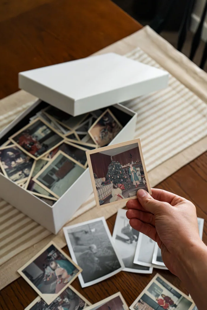 A hand holding a vintage family photo above a box of nostalgic family snapshots on a table.