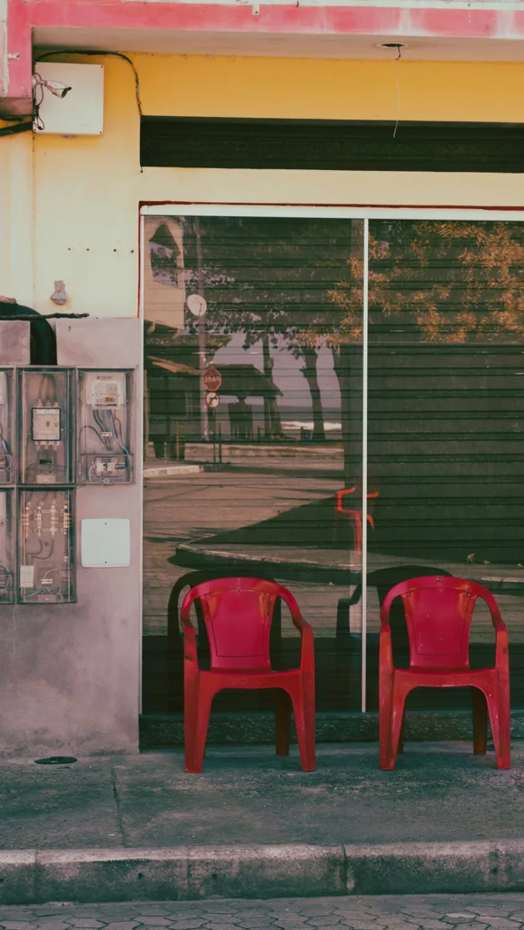 Two empty red chairs sit outside a closed shop with a glass window reflecting the street.