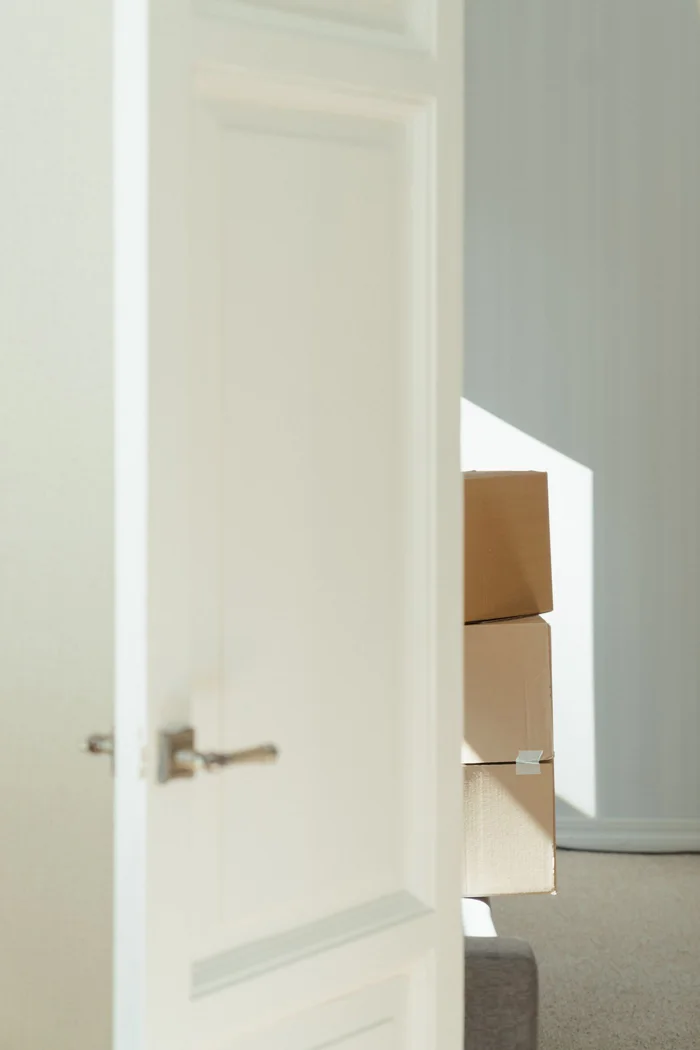 Open white door revealing stacked cardboard boxes in a bright, sunlit interior suggesting a moving or relocation scene.