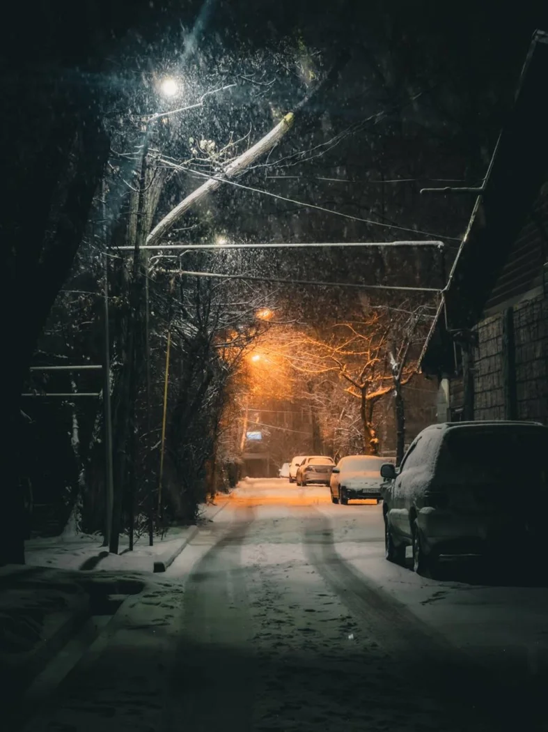 A quiet snowy night scene on a city street, with parked cars and glowing streetlights.