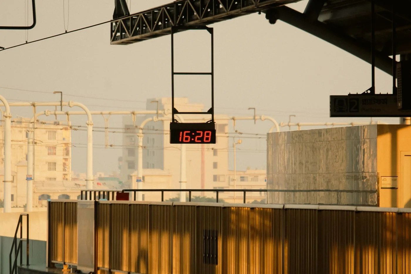 Digital clock displaying 16:28 at a railway station during golden hour, illuminated by warm sunset light.
