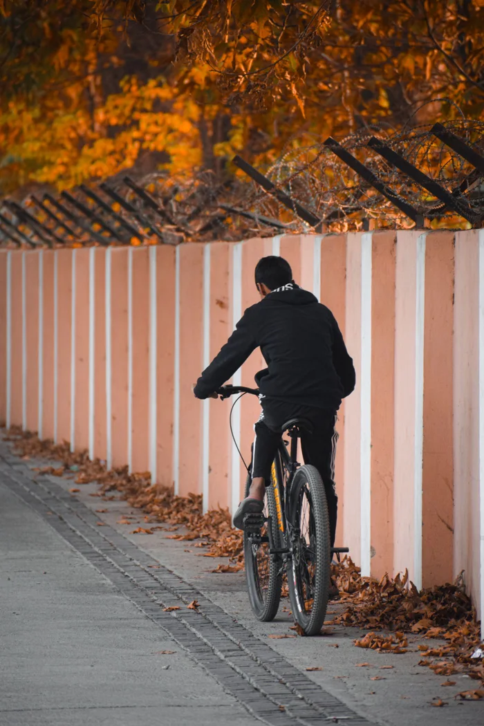 A teenager rides a bicycle along a leaf-lined pathway against an autumn wall backdrop.