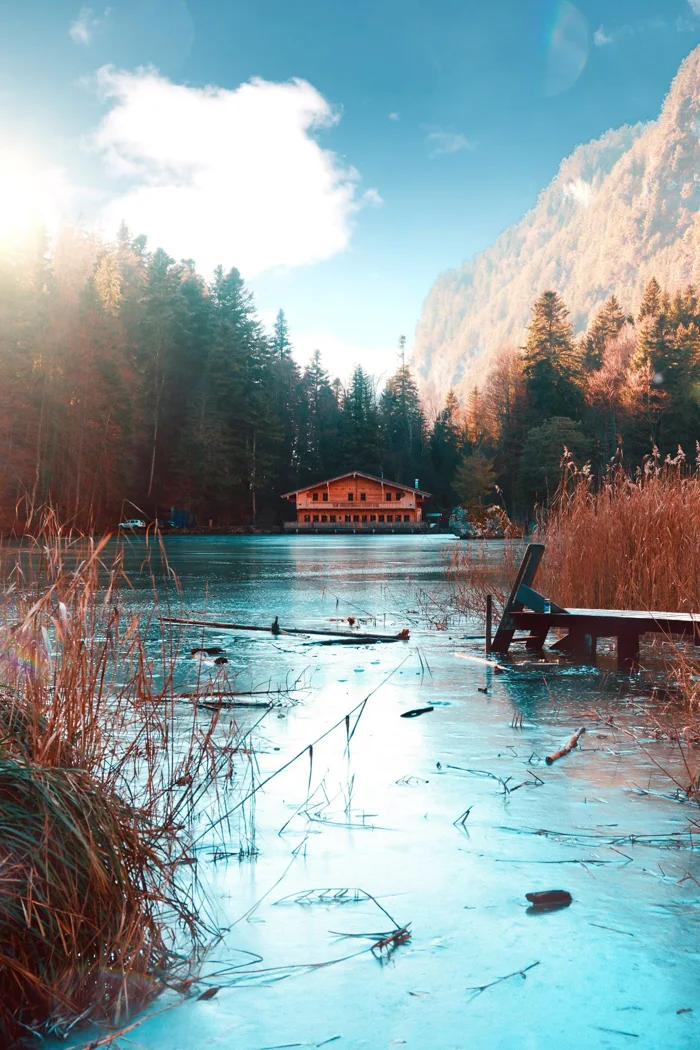 Beautiful winter scene of a cabin by a frozen lake with mountain backdrop under a bright sky.