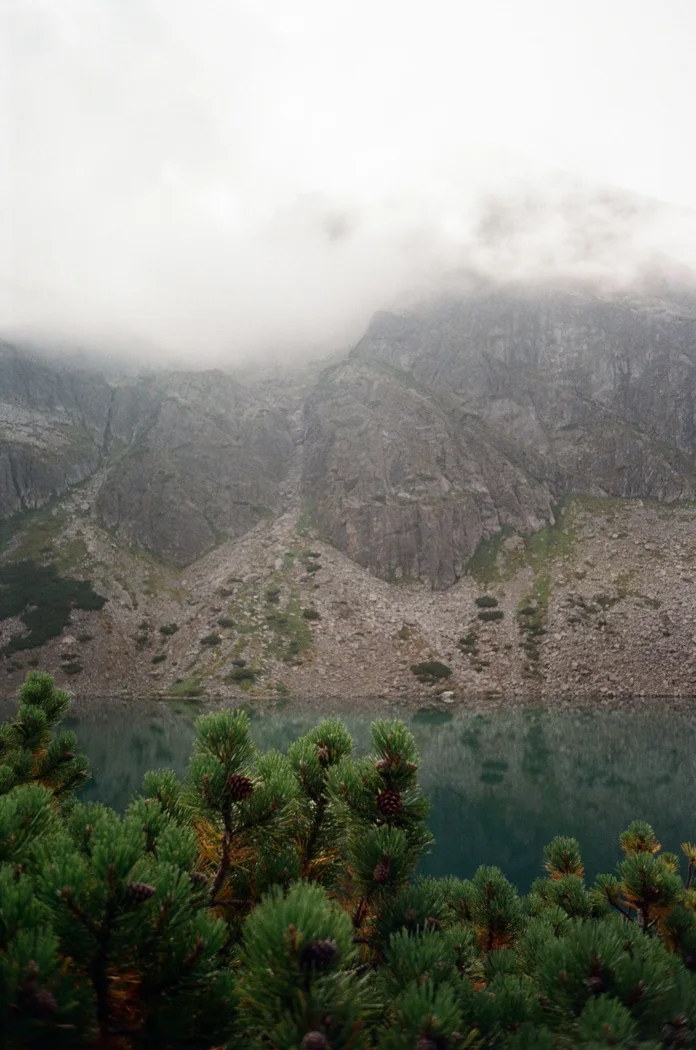 A serene mountain lake with misty clouds hovering over rocky cliffs, surrounded by lush green pine trees.