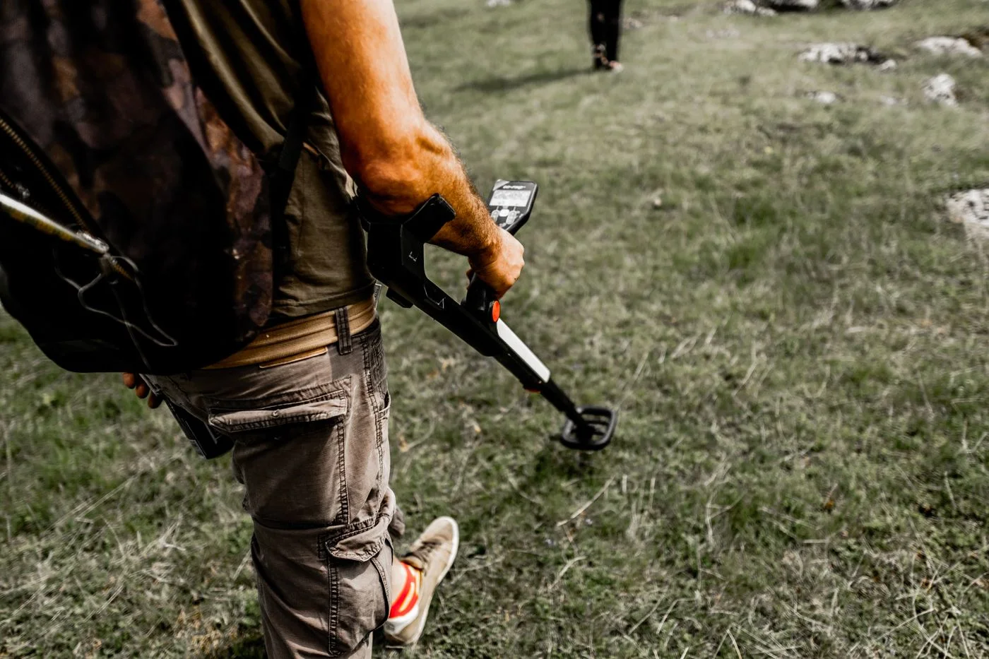 Person using a metal detector on a grassy field wearing casual attire.