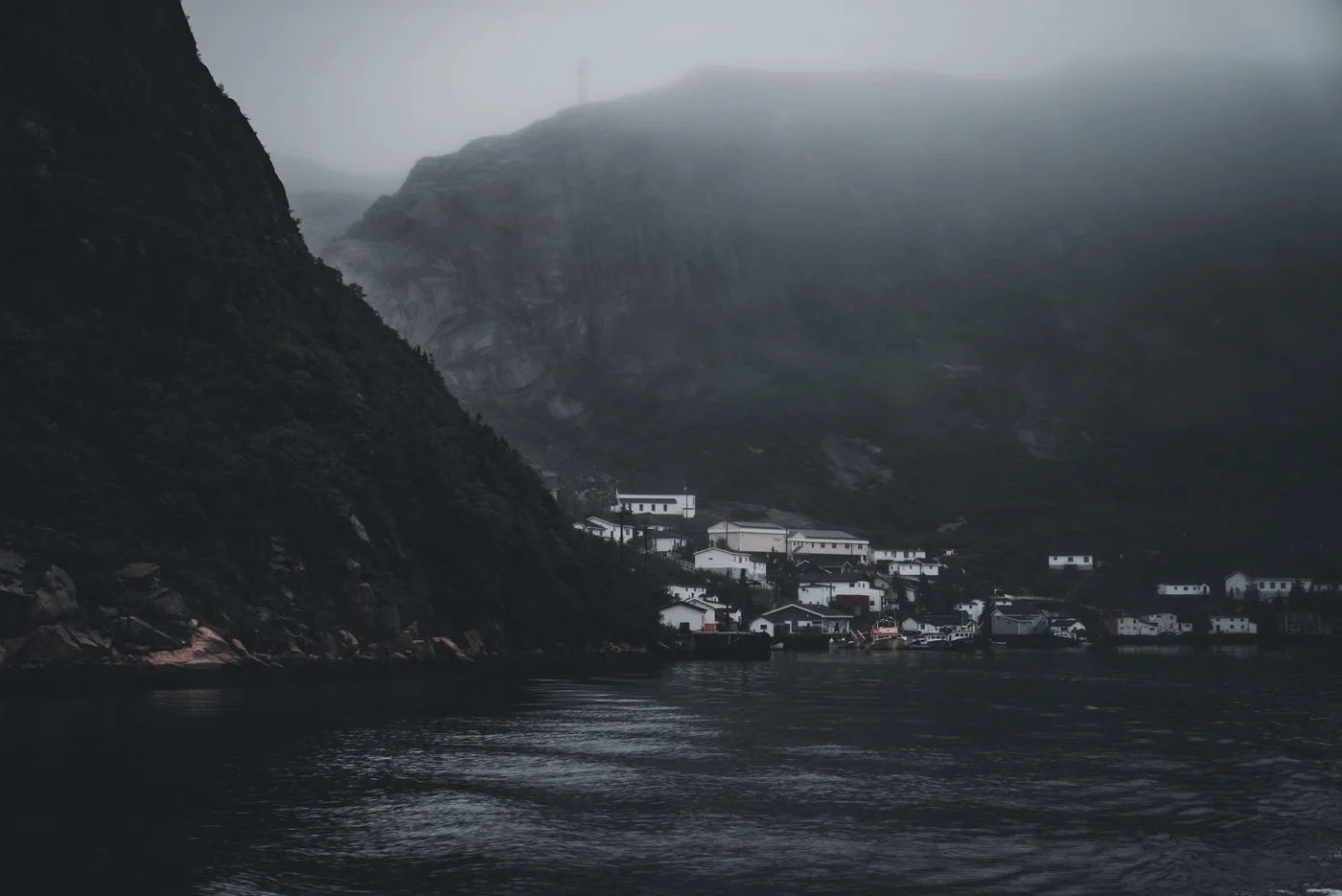 A foggy coastal village nestled under towering cliffs by the sea, creating a moody atmosphere.