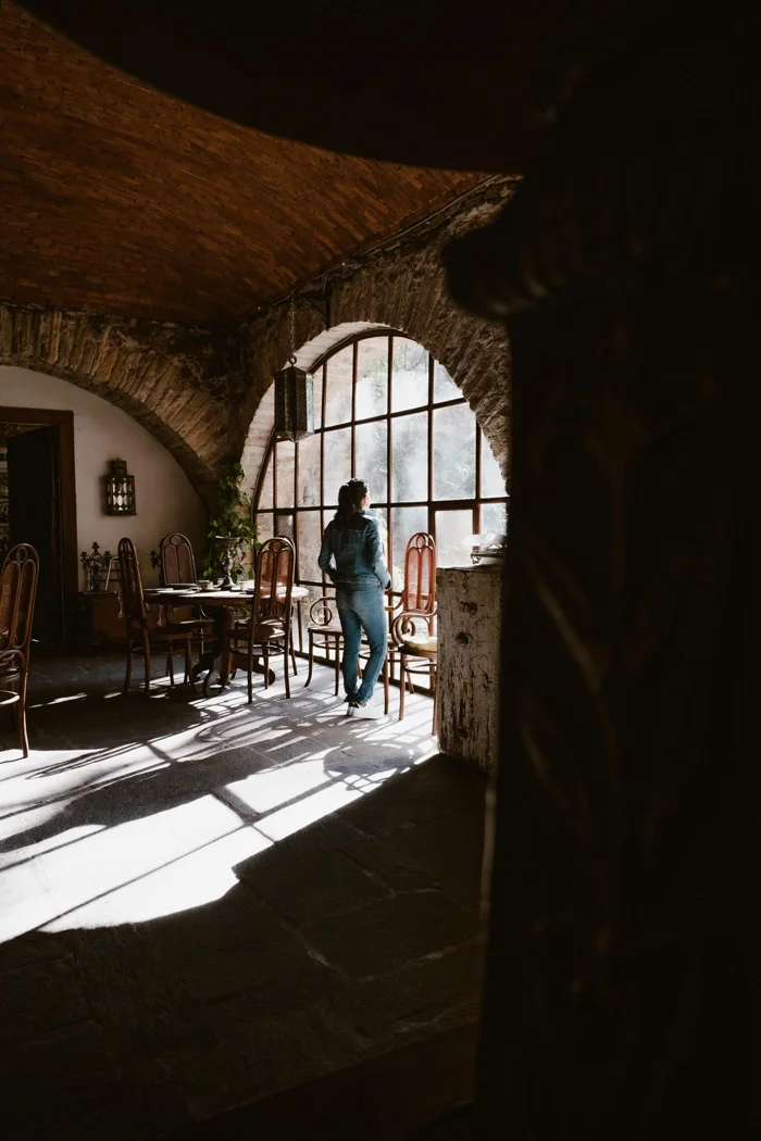 A woman stands gazing out a large arched window in a rustic, sunlit cafe with empty tables and chairs.