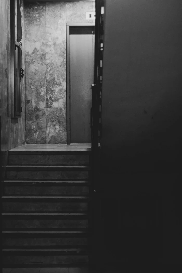 A black and white photograph of a dimly lit staircase leading to a closed door.