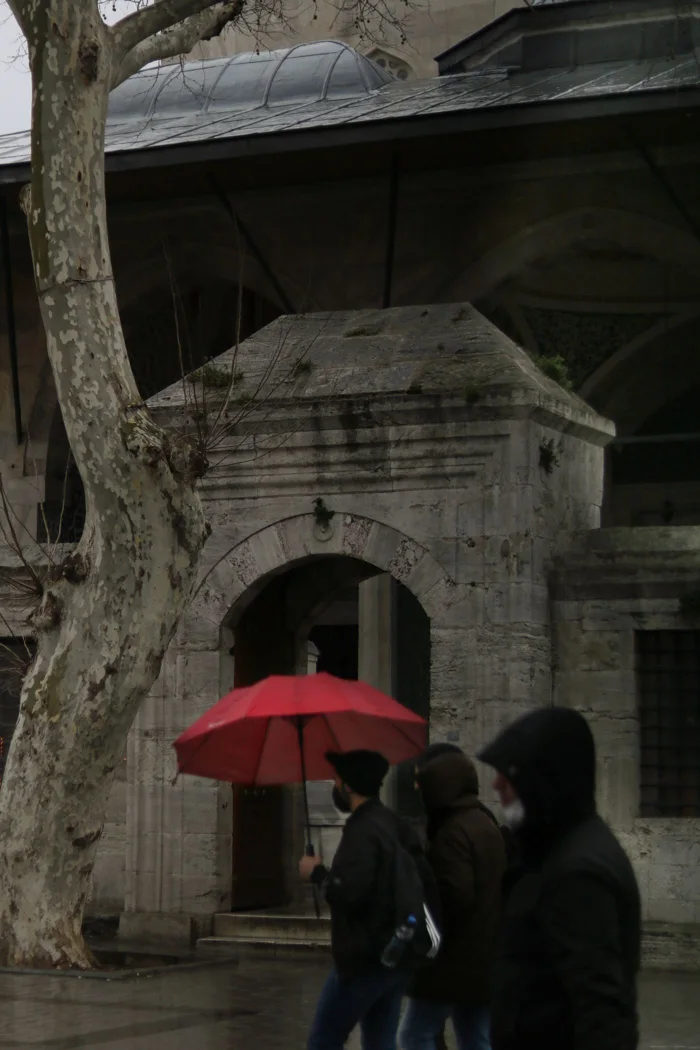 People walking with a red umbrella on a rainy urban street near a historic stone entrance.