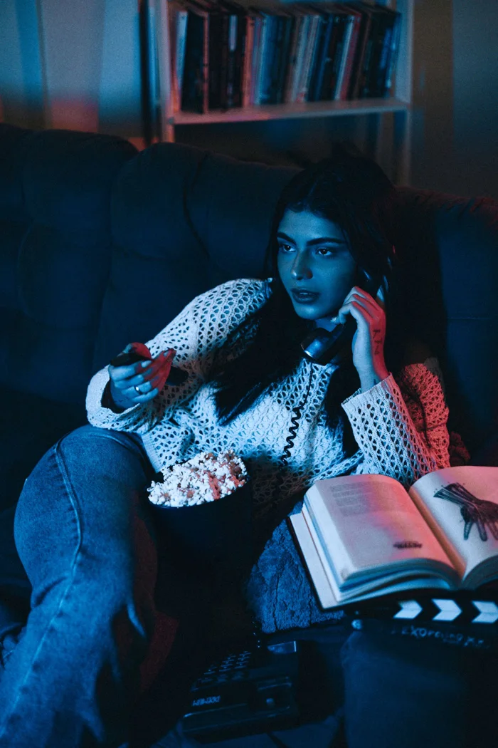 Young woman watching TV with remote, popcorn, and headphones in a cozy room setting.