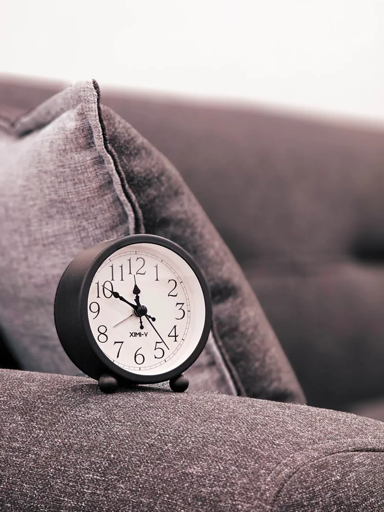 A black analog clock resting on a gray sofa with a cozy pillow, showcasing a minimalist home interior.