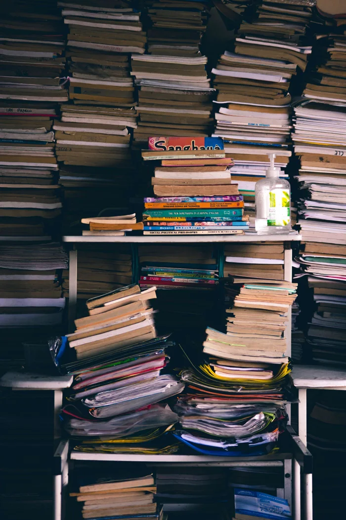 Full background of messy heaps of various paperback books stacked together on white shelves in light room.